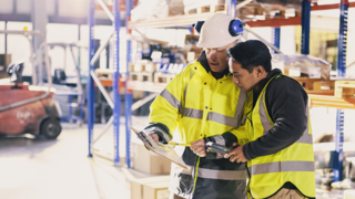 Two men wearing yellow jackets in a warehouse looking at a clipboard 